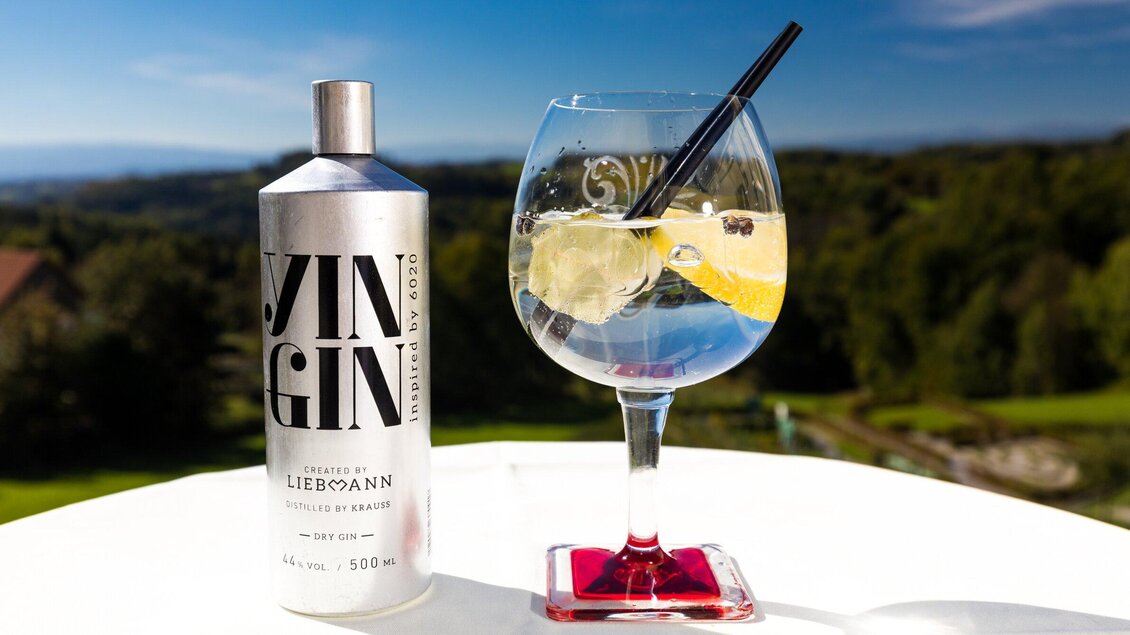 Ein erfrischendes Glas Gin mit Zitrone und Eiswürfeln steht auf einem Tisch. Im Hintergrund ist eine grüne Landschaft und ein blauer Himmel zu sehen. | © TV Region Graz - Harry Schiffer