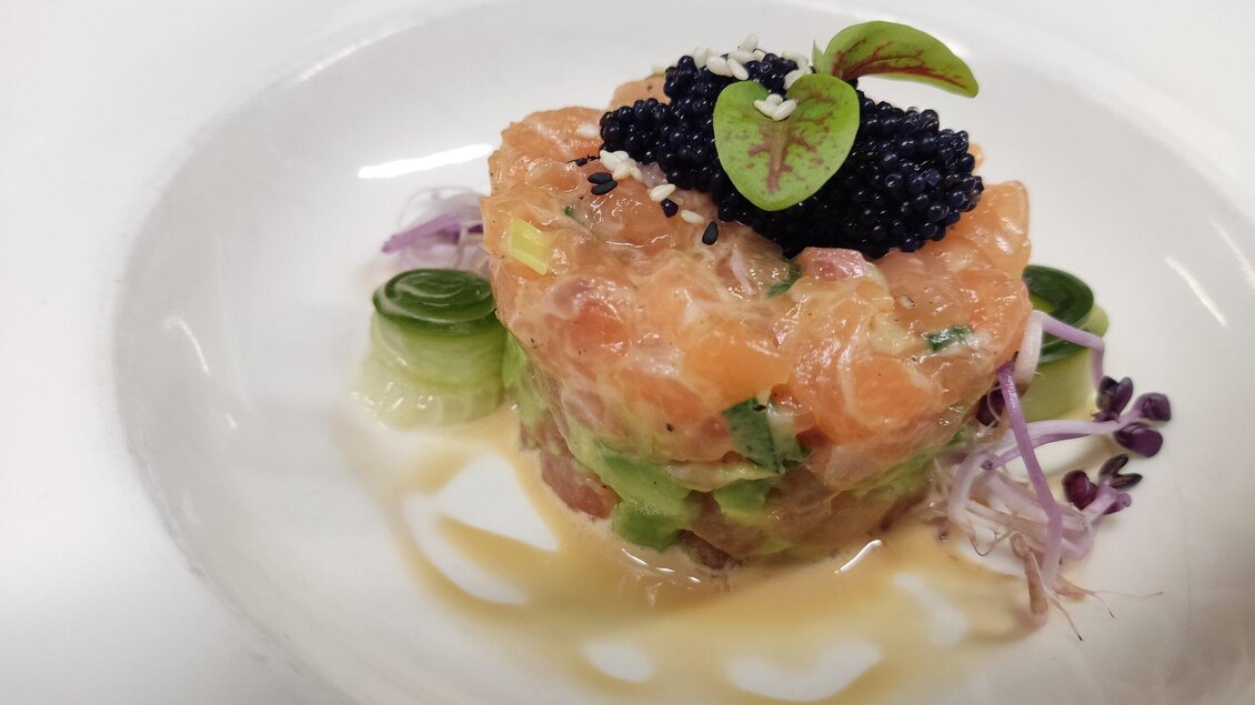 Ein kunstvoll angerichteter Lachstatar mit Avocado, garniert mit schwarzem Kaviar und frischen Kräutern. Das Gericht liegt auf einem weißen Teller und wird von zarten Pflanzen umrahmt. | © Hotel Liebmann