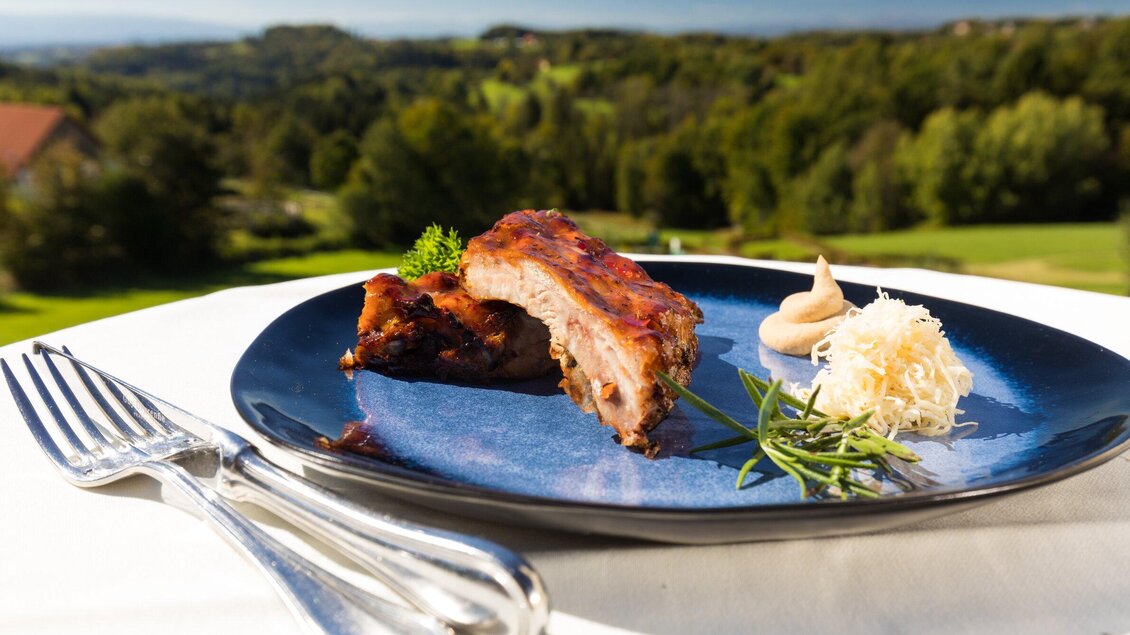 Ein schmackhaftes Stück Fleisch auf einem blauen Teller, garniert mit Meerrettich und frischen Kräutern. Im Hintergrund ist eine grüne Landschaft zu sehen. | © TV Region Graz - Harry Schiffer