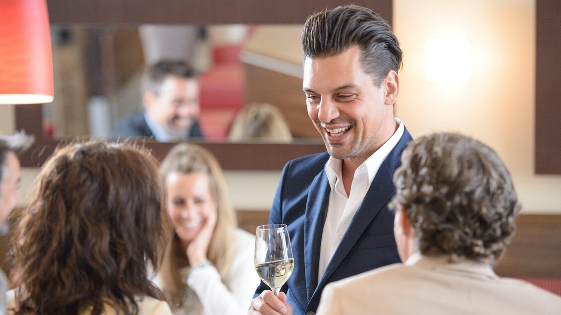 Ein lächelnder Kellner spricht mit Gästen in einem modernen Restaurant. Die Atmosphäre ist entspannt und einladend.