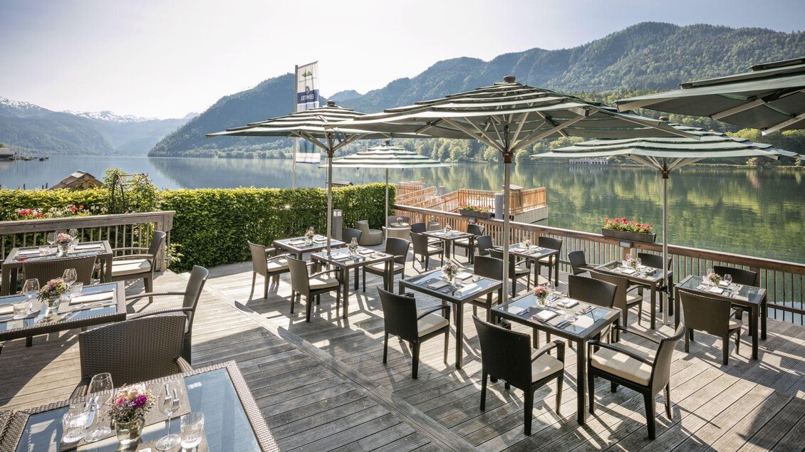 Ein einladendes Restaurant am See mit Terrassenstühlen und Tischen. Im Hintergrund sind Berge und ein klarer Himmel zu sehen. | © Seehotel Grundlsee