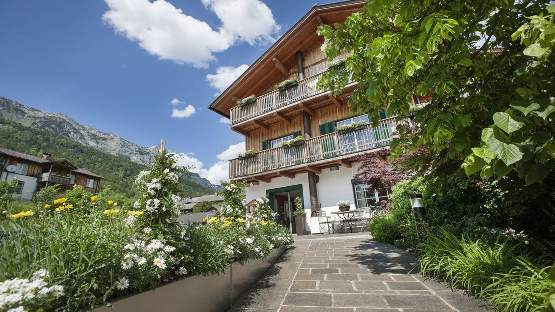 Ein charmantes Haus mit Holzverkleidung und Balkon, umgeben von bunten Blumen und grünen Pflanzen. Im Hintergrund sind Berge und ein bewölkter, blauer Himmel sichtbar. | © Seehotel Grundlsee