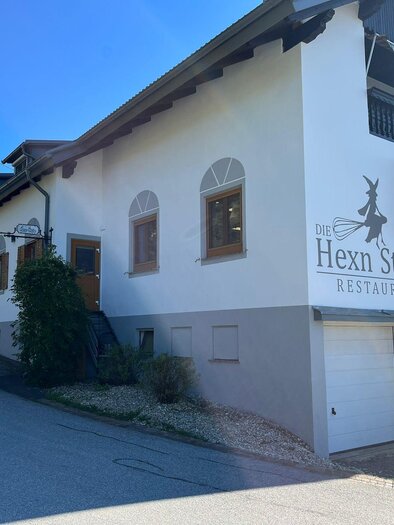 A rustic restaurant building named "Hexn Stubn" on the hillside. It has a bright facade and many windows, surrounded by a tranquil environment. | © Familie Wachmann