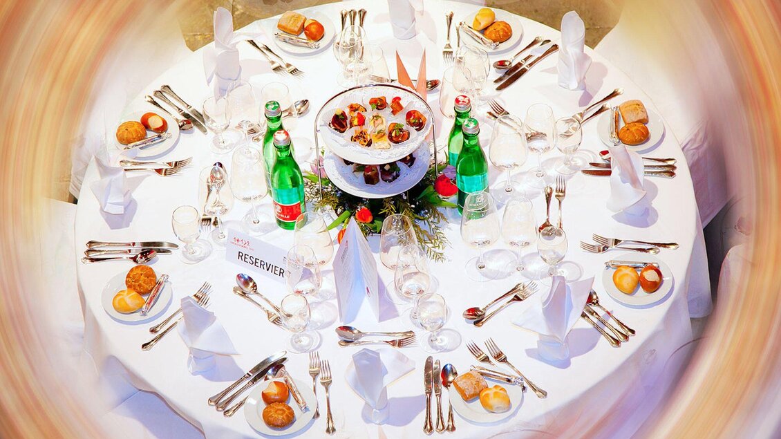 Ein schön gedeckter Tisch mit weißen Tischdecken und glänzendem Besteck. Auf dem Tisch stehen Wasserflaschen und ein zweistufiges Dessert-Tablett. | © Revita Gastronomie
