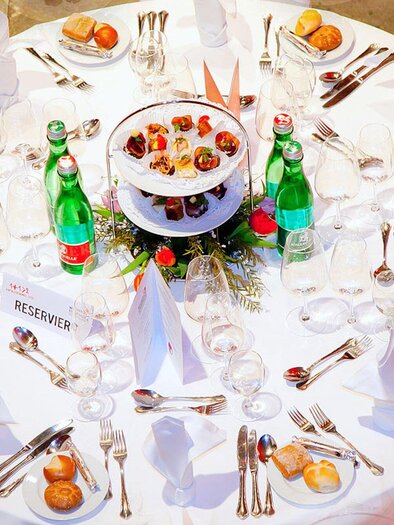 A beautifully set table with white tablecloths and shiny cutlery. On the table are water bottles and a two-tier dessert tray. | © Revita Gastronomie