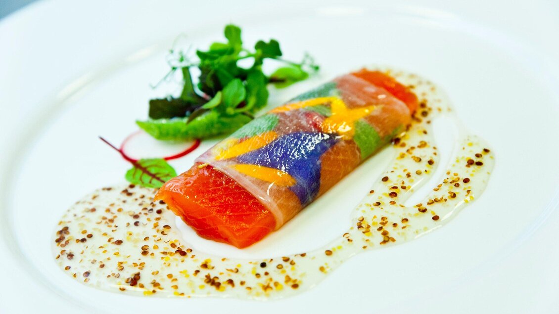 Ein kunstvoll angerichteter Teller mit Lachs, umhüllt von buntem Gemüse. Garniert mit frischen Kräutern und einer Sauce. | © Revita Gastronomie