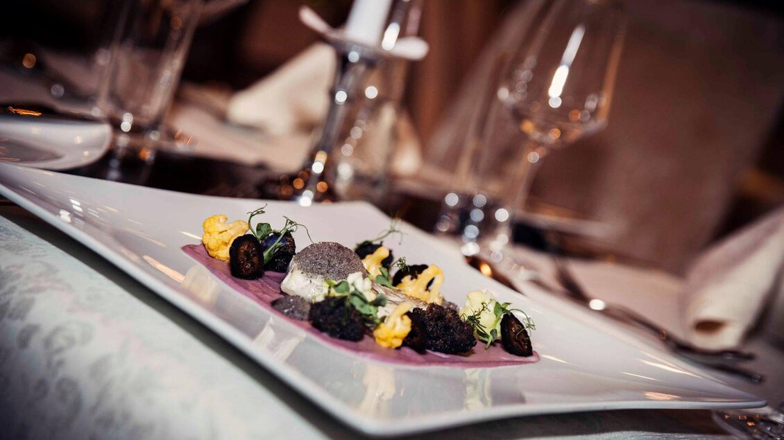 Ein elegant angerichteter Teller mit verschiedenen Köstlichkeiten, darunter bunte Garnierungen und feine Zutaten. Im Hintergrund sind Tischdekoration und Gläser sichtbar. | © Revita Gastronomie