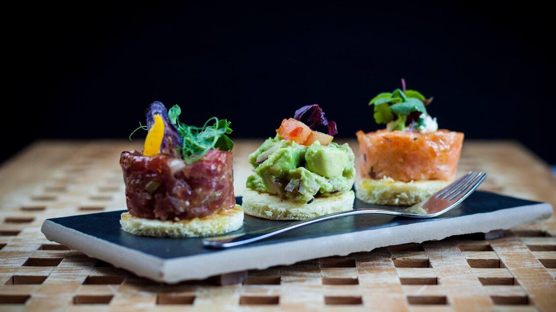Drei appetitliche Häppchen auf einem schwarzen Teller. Jedes Häppchen ist mit frischen Zutaten und Kräutern dekoriert. | © Revita Gastronomie