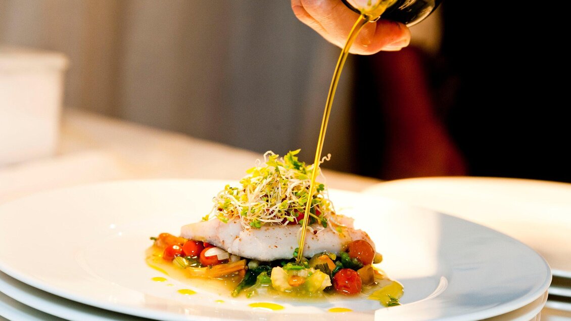 Ein kunstvoll angerichtetes Gericht mit Fisch, Gemüse und frischen Kräutern. Olivenöl wird gerade darüber gegossen. | © Revita Gastronomie