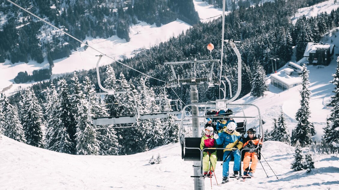 Eine Skiliftfahrt mit vier Fahrern in bunten Skianzügen. Im Hintergrund sind schneebedeckte Berge und grüne Tannenbäume zu sehen. | © Armin Walcher