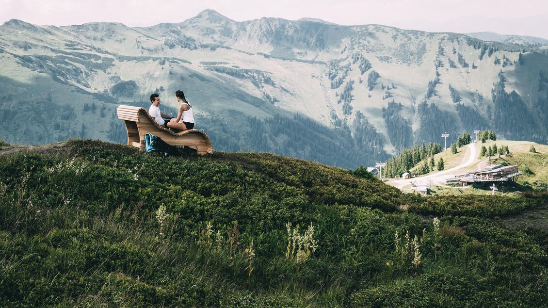 Zwei Personen sitzen auf einer Holzbank in der Natur. Im Hintergrund erstrecken sich grüne Berge und ein klarer Himmel. | © Armin Walcher