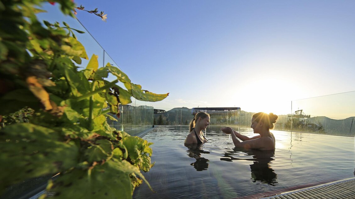 Zwei Frauen entspannen in einem Infinity-Pool bei Sonnenuntergang. Die Umgebung ist grün und einladend, mit Pflanzen im Vordergrund. | © Herbert Raffalt