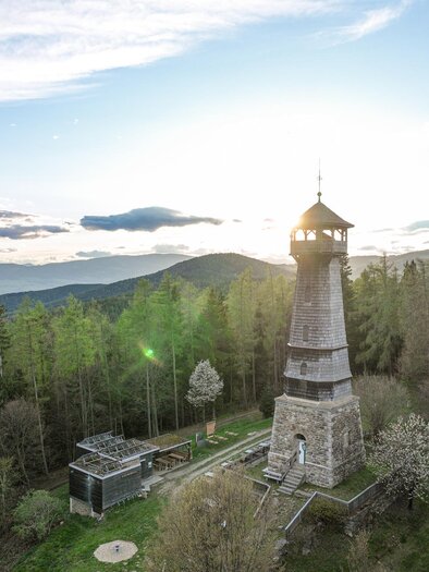 Ein Aussichtsturm inmitten eines Waldes mit sanften Hügeln im Hintergrund. Die Sonne geht hinter den Bergen unter und sorgt für eine malerische Stimmung. | © Oststeiermark Tourismus