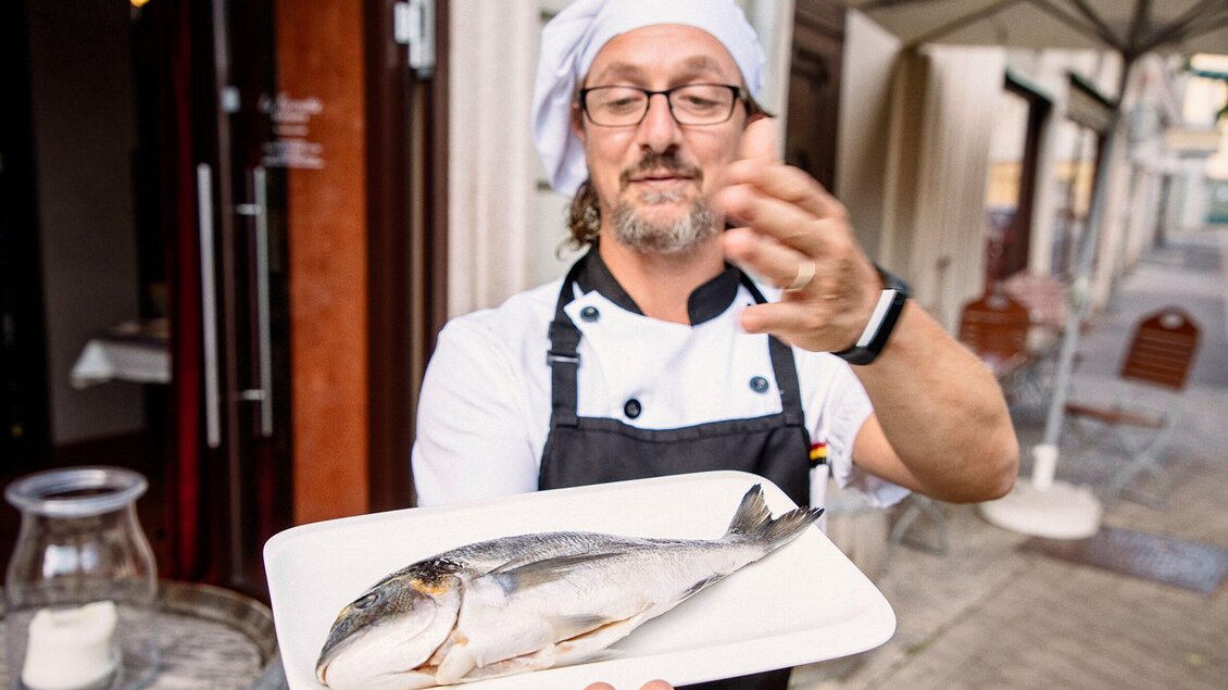 Ein Koch präsentiert stolz einen frischen Fisch auf einem Teller. Im Hintergrund ist eine gemütliche Straßenatmosphäre zu sehen. | © La Tavernetta