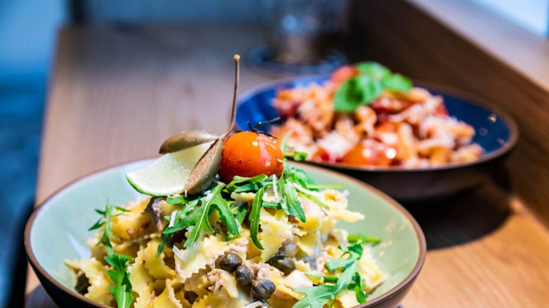 Ein Teller mit Pasta, garniert mit Rucola, Kapern und einer Kirschtomate. Im Hintergrund befindet sich ein weiterer Teller mit einer anderen Pasta-Variation. | © Ristorante Trenta