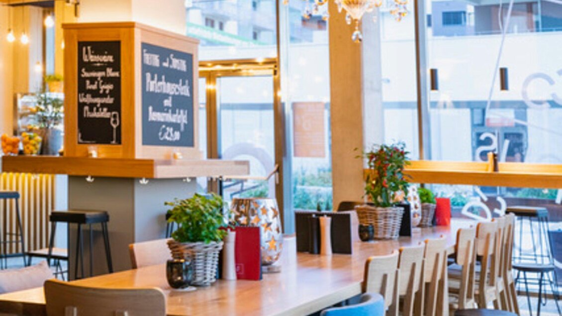 Ein modernes Restaurant mit einem langen Holztisch und Stühlen. Große Fenster lassen viel Licht herein und schaffen eine einladende Atmosphäre. | © Ristorante Trenta