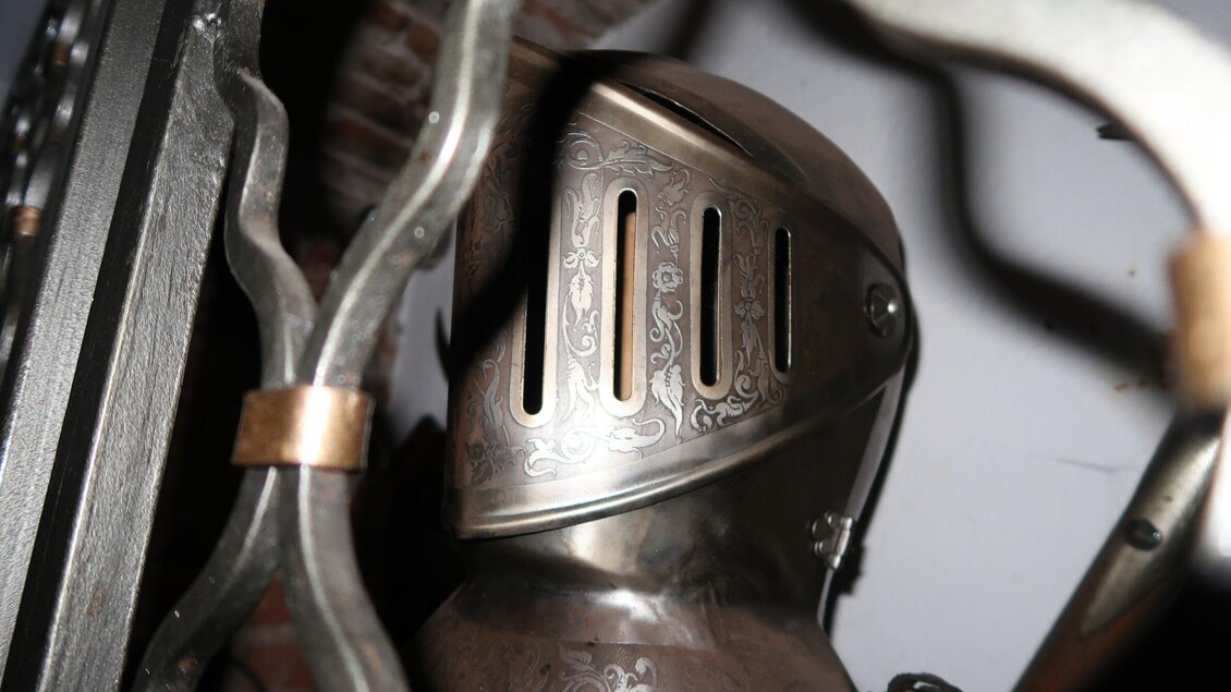 Ein glänzender Ritterhelm mit feinen Gravuren. Im Hintergrund sind Teile von Rüstungen sichtbar. | © Ritterwirt