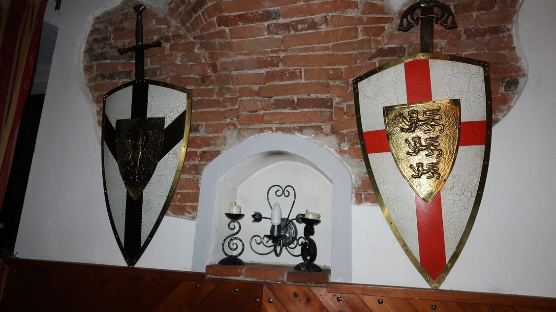 Zwei dekorative Ritterrüstungen hängen an einer Backsteinwand. Links ist ein schwarzes Schild und rechts ein weißes Schild mit rotem Kreuz. | © Ritterwirt