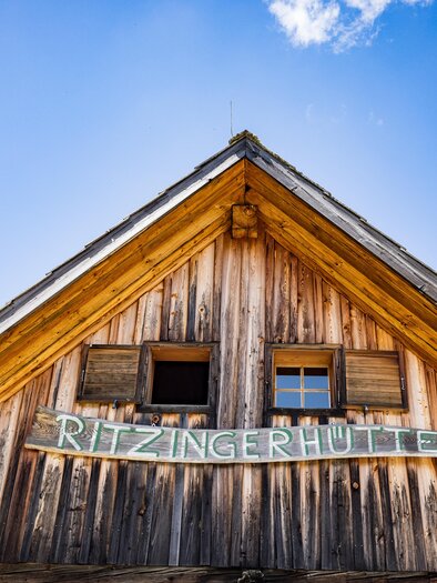 A traditional wooden house with a pointed roof. Above the entrance, the inscription "Ritzingerhütte" is displayed. | © Netzwerk Kulinarik Wildbild