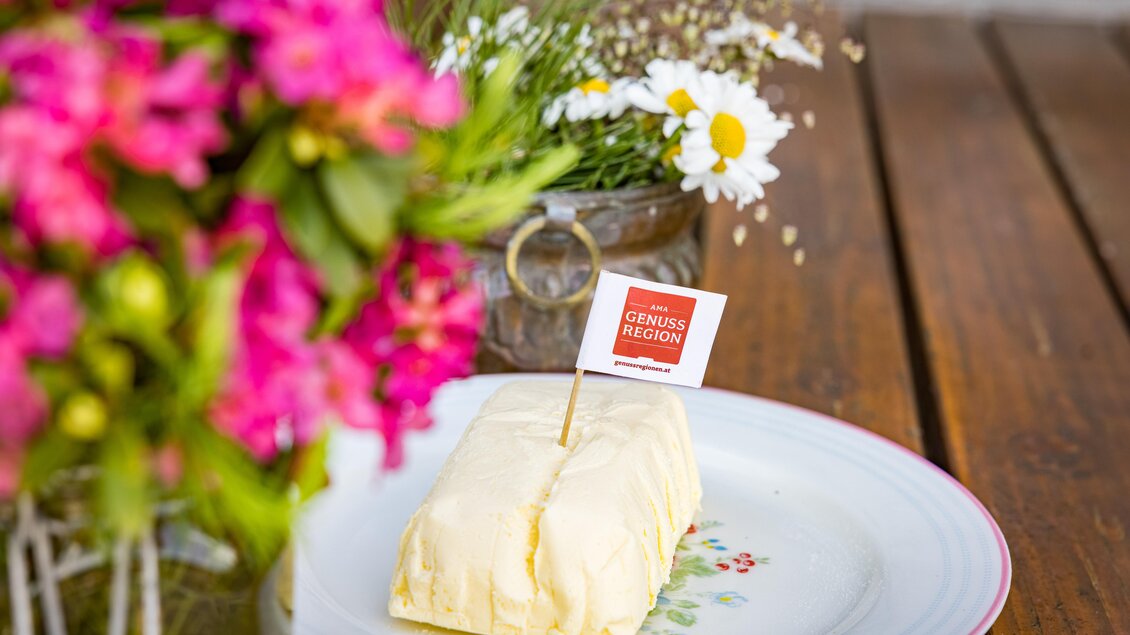 Ein Stück Butter auf einem blumendekorierten Teller mit einem kleinen Fähnchen. Im Hintergrund sind bunte Blumen und eine rustikale Holzoberfläche zu sehen. | © Netzwerk Kulinarik Wildbild