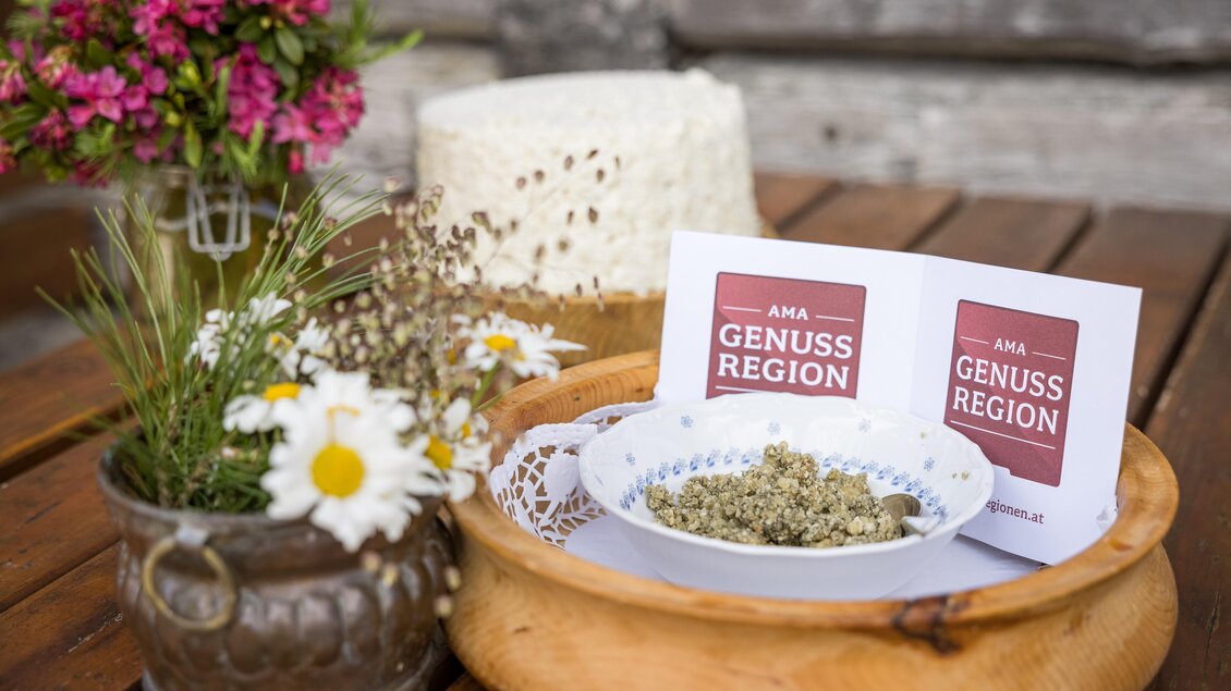 Ein rustikales Arrangement mit einer Holzschale, in der Kräuter liegen. Dazu gehören eine kleine Blumenvase und zwei Karten mit dem Aufdruck „AMA GENUSS REGION“. | © Netzwerk Kulinarik Wildbild