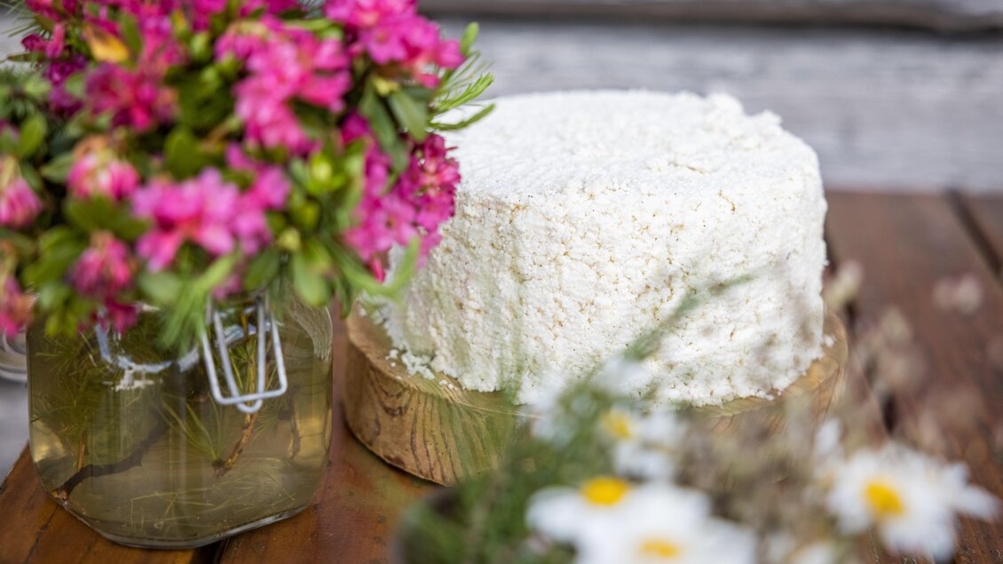 Ein runder, weißer Kuchen steht auf einem Holzbrett. Daneben befindet sich ein Glas mit bunten Blumen. | © Netzwerk Kulinarik Wildbild