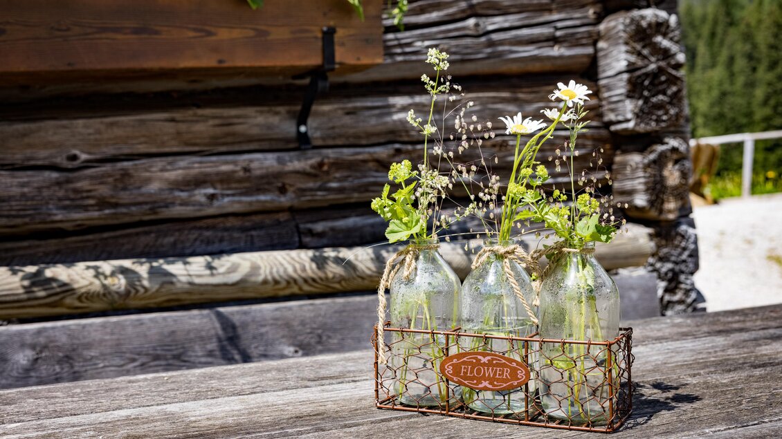 Drei Glasflaschen mit frischen Blumen stehen in einem Drahtkorb auf einem Holztisch. Im Hintergrund sieht man eine rustikale Holzstruktur und grüne Bäume. | © Netzwerk Kulinarik Wildbild