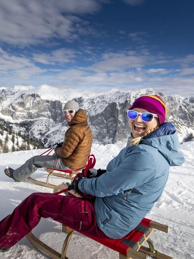 Zwei Menschen sitzen auf Schlitten im Schnee und lachen. Im Hintergrund sind schneebedeckte Berge und ein blauer Himmel zu sehen. | © TVB Ausseerland - Salzkammergut-Tom Lamm