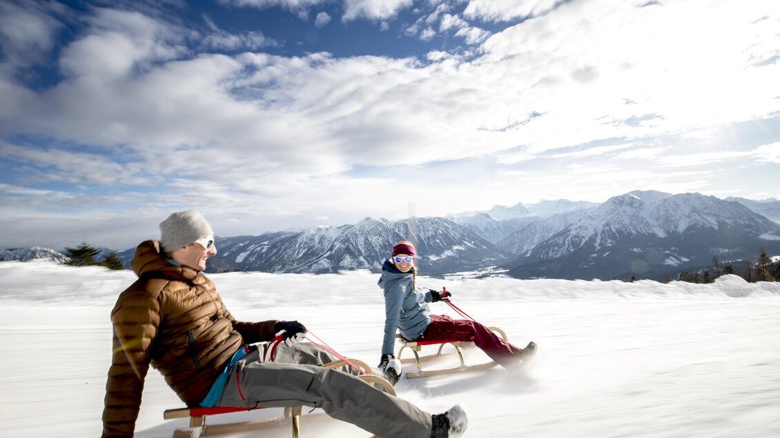 Zwei Personen fahren mit dem Schlitten im Schnee. Im Hintergrund sind majestätische Berge und ein blauer Himmel zu sehen. | © TVB Ausseerland - Salzkammergut/Tom Lamm