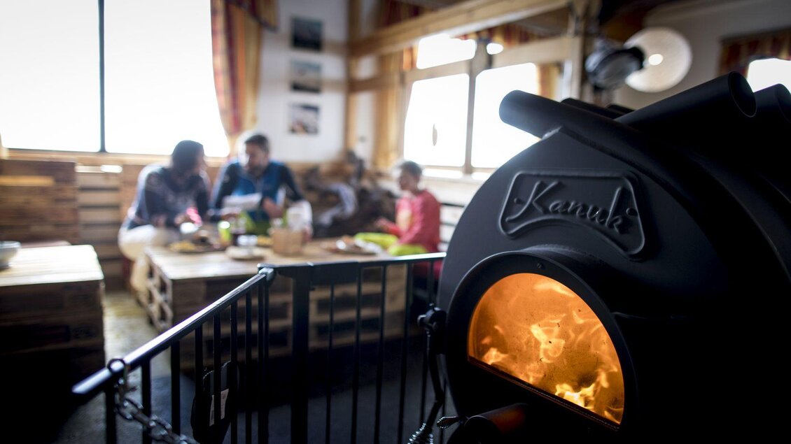 Ein gemütlicher Wohnraum mit einem Holzofen im Vordergrund. Im Hintergrund sitzen drei Menschen an einem Tisch und genießen eine Mahlzeit. | © TVB Ausseerland - Salzkammergut-Tom Lamm