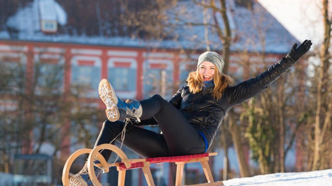 Eine fröhliche Frau sitzt auf einem Schlitten und genießt das Winterwetter. Im Hintergrund sind schneebedeckte Bäume und eine historische Gebäudeansicht zu sehen. | © TV Region Graz-MiasPhotoart
