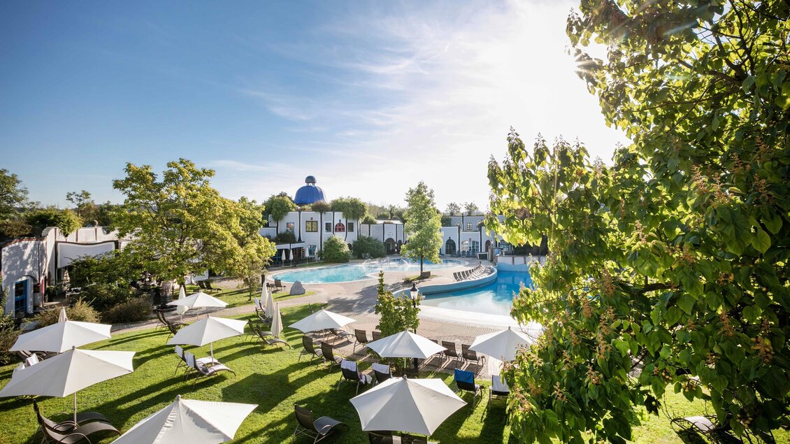 Eine gemütliche Poollandschaft umgeben von grünen Bäumen und Liegen. Weiße Sonnenschirme bieten Schatten für Entspannung im Freien. | © Rogner Bad Blumau (c) Hundertwasser Architekturprojekt