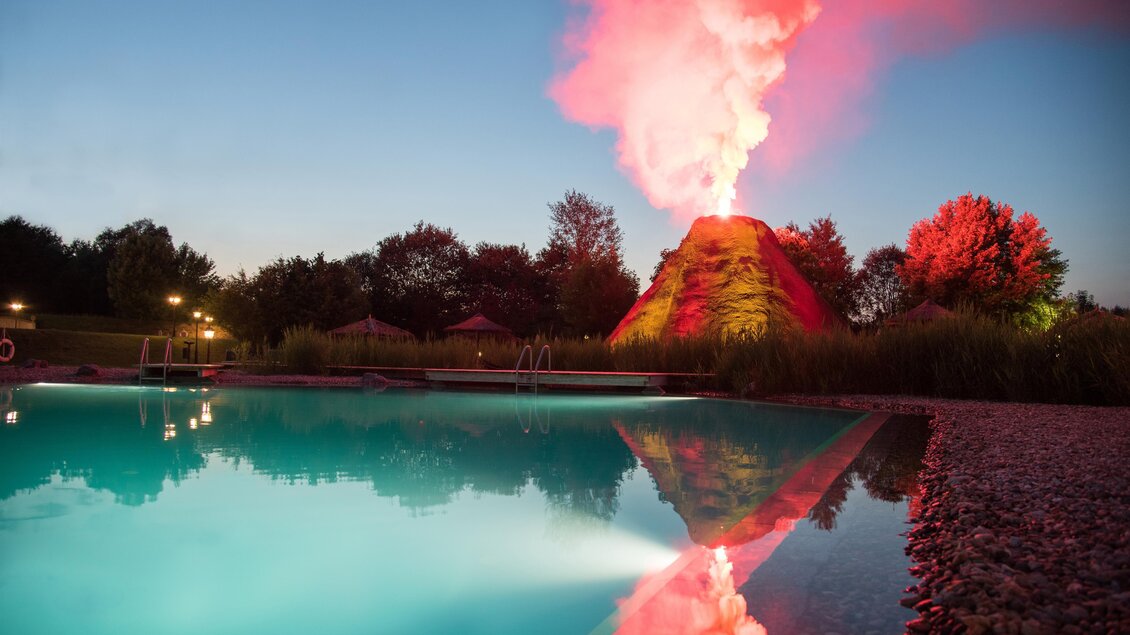 Ein künstlicher Vulkan spuckt bunten Rauch in den Abendhimmel. Im Vordergrund sieht man einen klaren Pool, der die Umgebung spiegelt. | © Rogner Bad Blumau