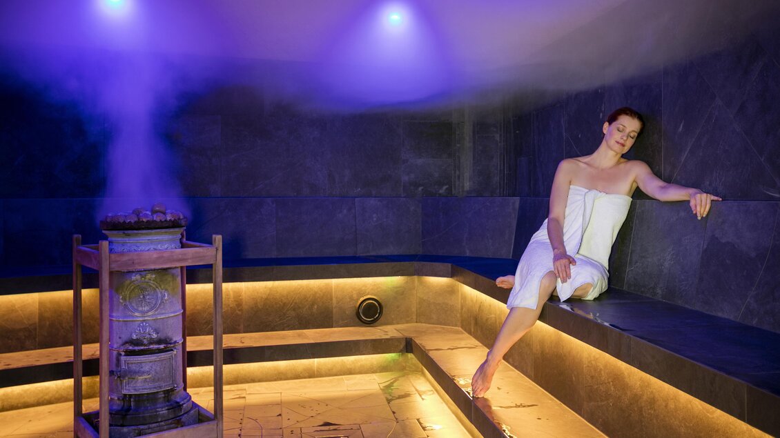 Ein entspannender Saunabereich mit sanftem Licht und Dampfkessel. Eine Frau sitzt entspannt auf einer Bank in der Sauna. | © Rogner Bad Blumau © Hundertwasser Architekturprojekt