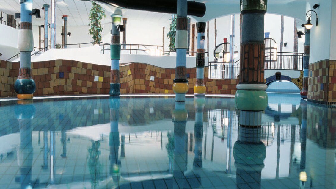 Ein stilvolles Hallenbad mit bunten Säulen und klaren Wasserflächen. Die farbenfrohen Fliesen und das natürliche Licht schaffen eine einladende Atmosphäre. | © Rogner Bad Blumau/Hundertwasser Architekturprojekt