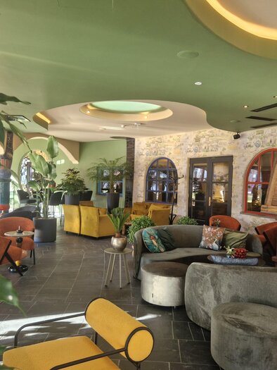 A modern lounge with cozy seating areas and an inviting atmosphere. Large windows and plants give the space a fresh ambiance. | © Rogner Bad Blumau/Hundertwasser Architekturprojekt