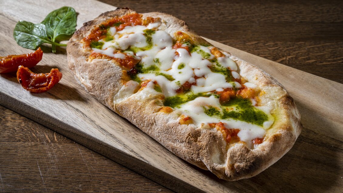 Ein frisch gebackenes Fladenbrot mit Tomatensoße, Käse und Pesto auf einem Holzbrett. Neben dem Fladenbrot liegen einige sonngetrocknete Tomaten und frische Basilikumblätter. | © Rogner Bad Blumau