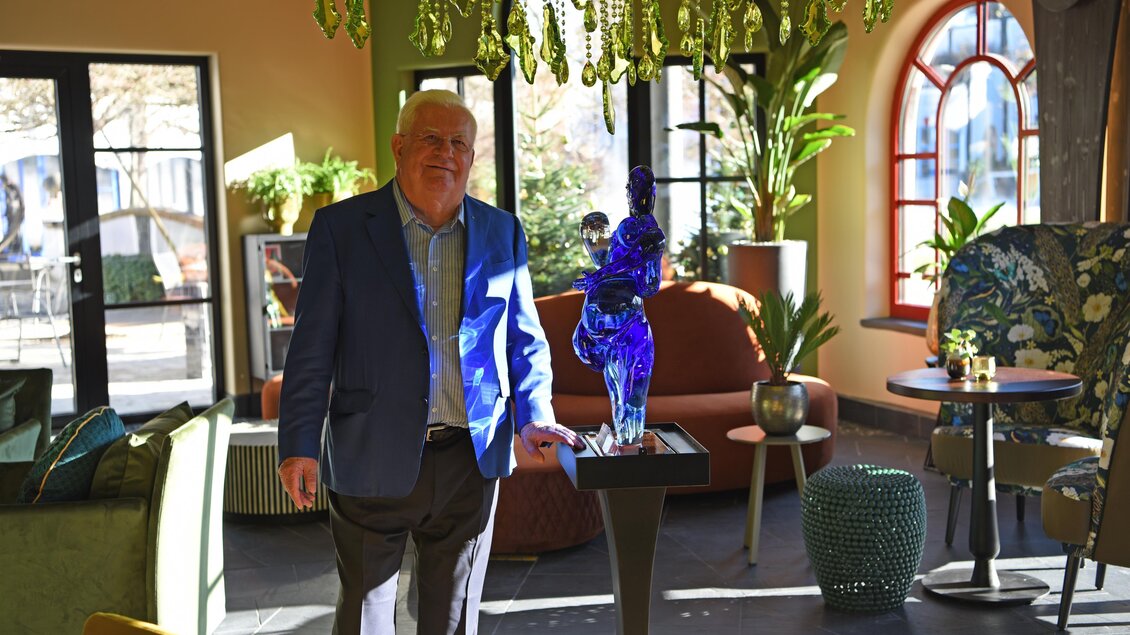Ein elegantes Wohnzimmer mit modernen Möbeln und einer bunten Dekoration. Eine Person steht neben einer blauen Skulptur auf einem Tisch. | © Rogner Bad Blumau/Hundertwasser Architekturprojekt