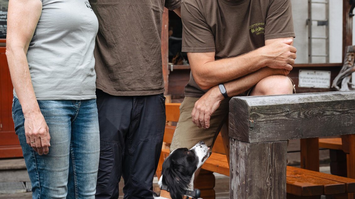 Eine Gruppe von drei Personen steht vor einer Hütte. Ein Hund sitzt auf dem Boden und schaut freundlich in die Kamera. | © Christoph Lukas