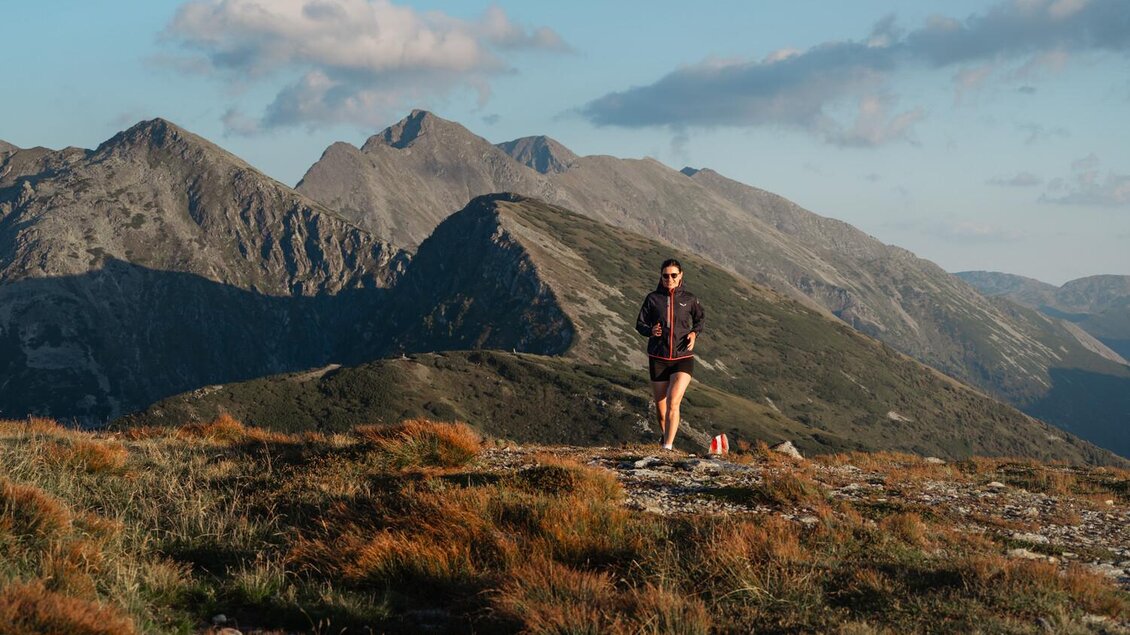 Eine Person läuft auf einem Berghang mit beeindruckenden Felsen und Bergen im Hintergrund. Die Landschaft ist grün und sonnig, perfekt für Outdoor-Aktivitäten. | © Christoph Lukas