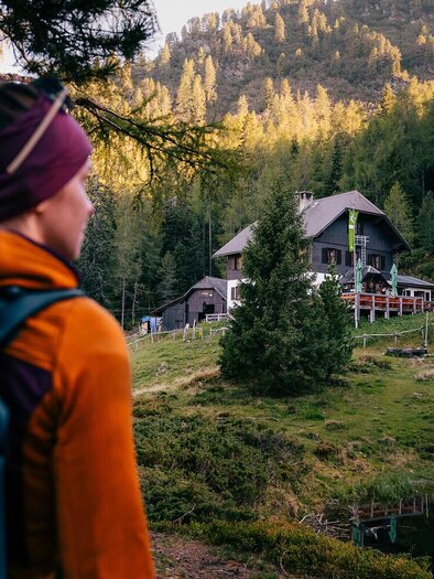 A hiker with a backpack looks at a mountain cabin nestled among trees. In the background, gentle hills and a clear sky can be seen.
