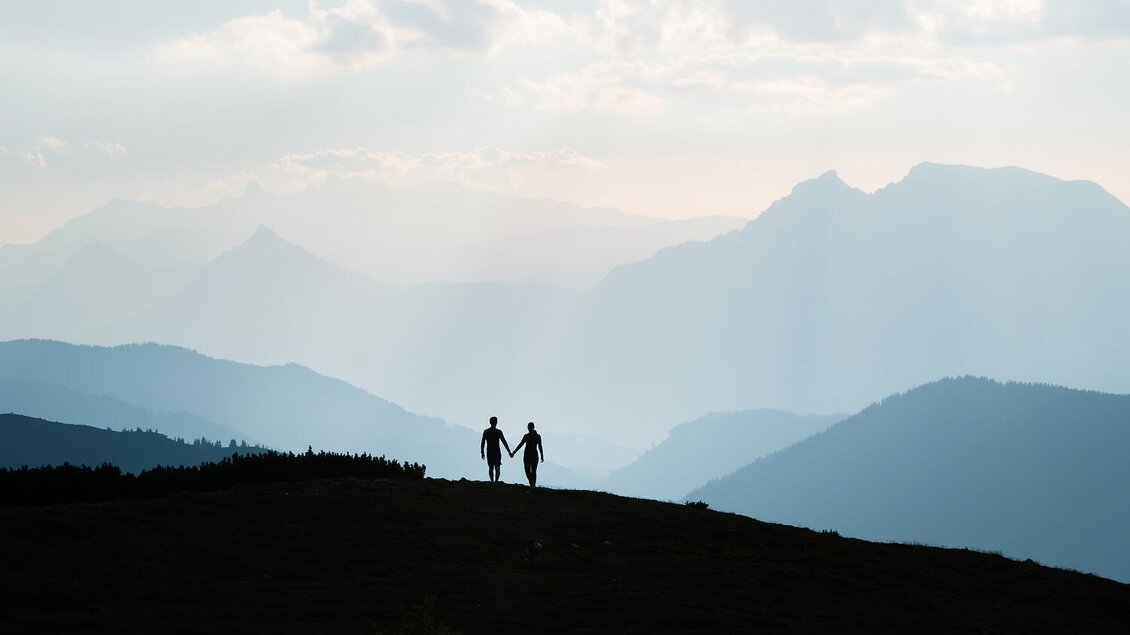 Ein Paar hält Händchen und steht auf einem Hügel mit einer beeindruckenden Berglandschaft im Hintergrund. Der Himmel ist bewölkt und strahlt eine ruhige Stimmung aus.