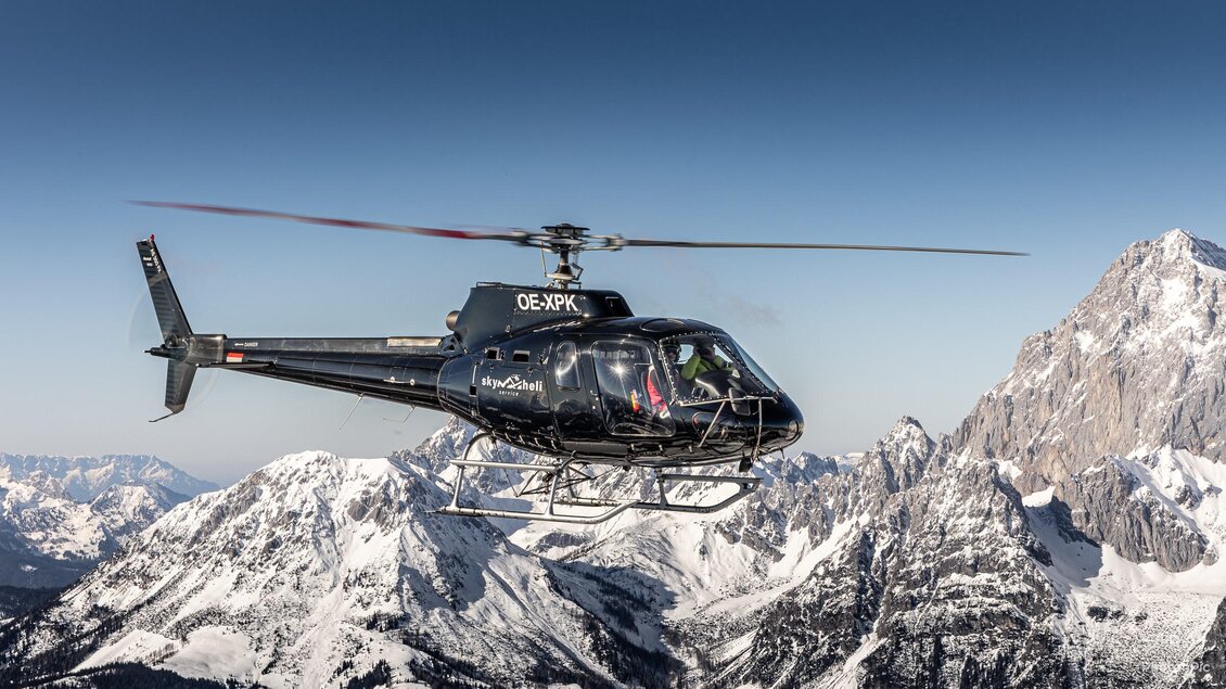Ein schwarzer Hubschrauber schwebt über schneebedeckten Bergen. Der Himmel ist klar und blau.