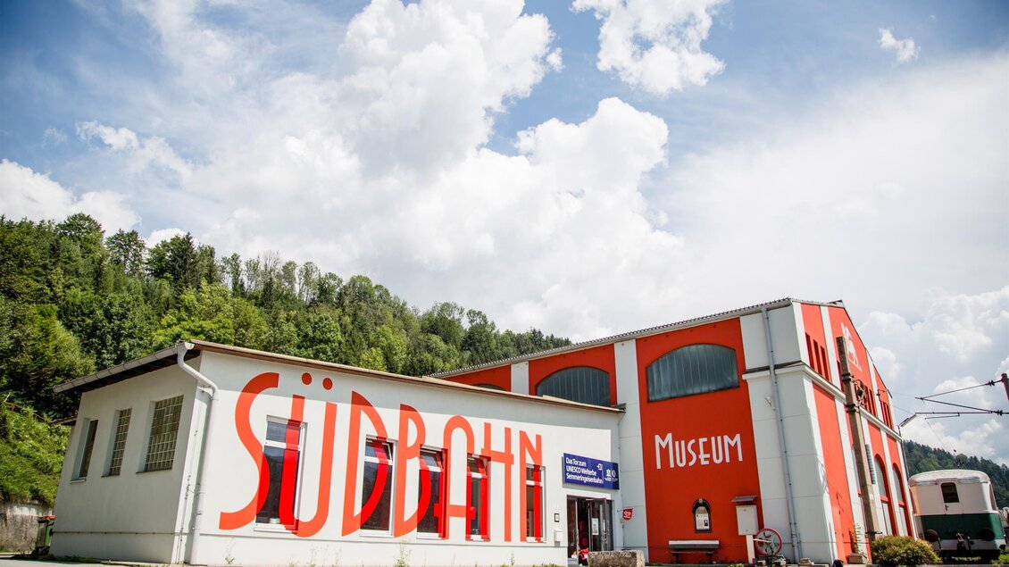 Ein buntes Gebäude mit dem Schriftzug „Südbahn Museum“ an der Fassade. Im Hintergrund sind Wälder und ein blauer Himmel mit weißen Wolken sichtbar. | © Südbahnmuseum