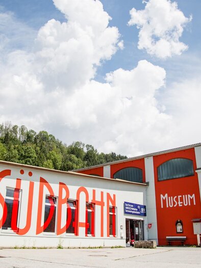 A colorful building with the inscription "Südbahn Museum" on the facade. In the background, forests and a blue sky with white clouds are visible. | © Südbahnmuseum