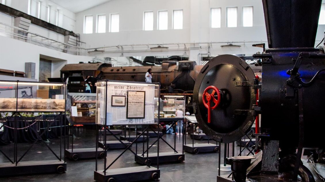 Ein modernes Museum mit historischen Dampflokomotiven. Im Vordergrund ist eine Lokomotive und im Hintergrund sind Vitrinen mit weiteren Exponaten zu sehen. | © Südbahnmuseum
