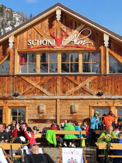 A rustic mountain cabin with a wooden façade and many guests outdoors. Surrounded by snow-capped mountains, the visitors enjoy the nice weather. | © Schöni Alm