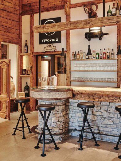 A cozy bar with wooden decor and a stone wall. The counter is equipped with several seats, and behind the bar, there are various drinks. | © Felix Werinos