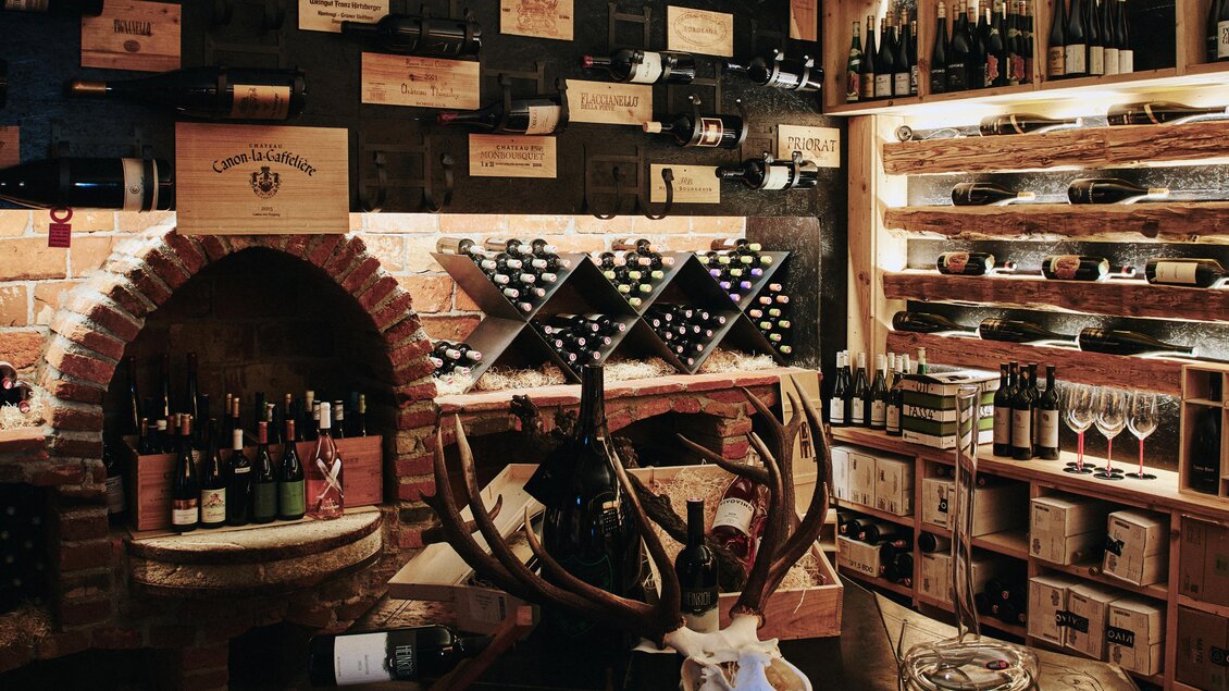 Ein gemütlicher Weinkeller mit Regalen voller Weinflaschen. An der Wand sind Holzboxen und ein dekorativer Geweihkopf zu sehen. | © Felix Werinos