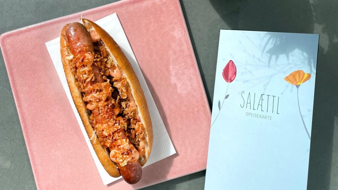 Ein Hotdog mit zarter Wurst und knusprigem Belag liegt auf einem rosa Teller. Daneben befindet sich ein Menü mit bunten Blumenmotiven. | © Salaettl